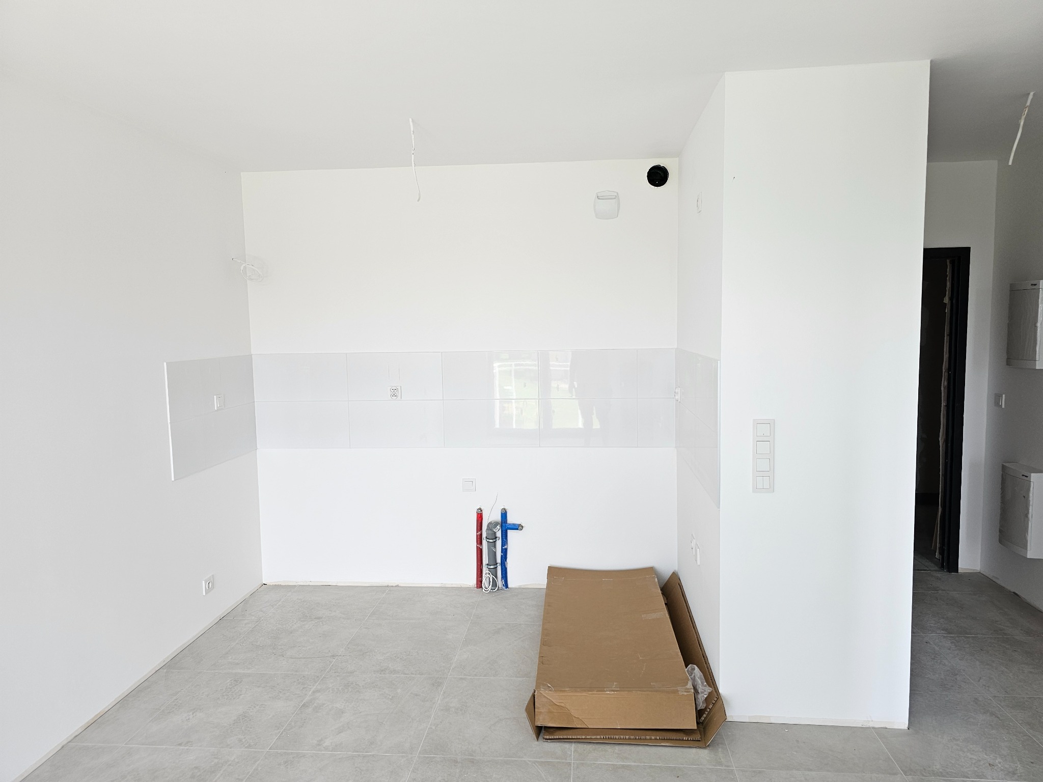 Białe wnętrze kuchni w trakcie wykańczania: ułożone płytki na ścianie, wyprowadzone instalacje wodne, szare płytki podłogowe, kartony na podłodze, widok na sąsiednie pomieszczenie.