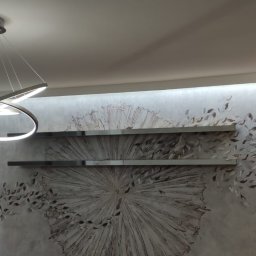 Usługi remontowo budowlane Pablobud Paweł Janowski - Nowoczesne oświetlenie sufitowe w kształcie pierścieni nad ścianą z dekoracyjną fakturą i metalowymi półkami.