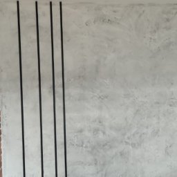 Usługi remontowo budowlane Pablobud Paweł Janowski - Ściana z efektem betonu architektonicznego, ozdobnymi czarnymi listwami i fragmentami muru z cegły, widoczna drabina malarska w tle.