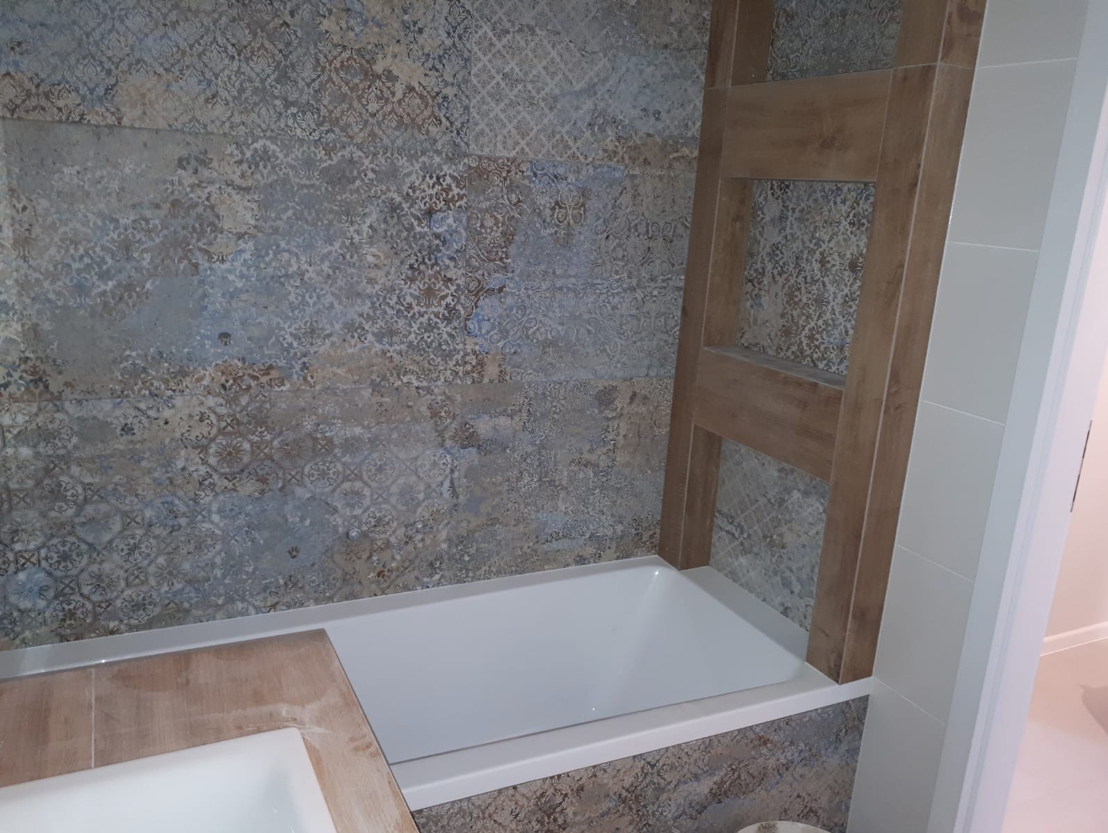 Wykończona łazienka z wanną obudowaną płytkami imitującymi drewno, ściany wyłożone płytkami w stylu vintage z motywami roślinnymi, drewniany stelaż na ręczniki.