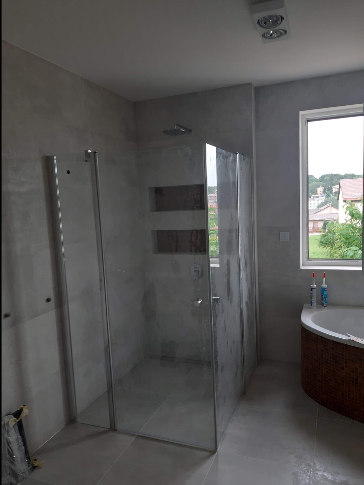 Wykończona łazienka z kabiną prysznicową typu walk-in, szare ściany z wnękami, okrągła wanna z mozaiką i widok na zieleń za oknem.