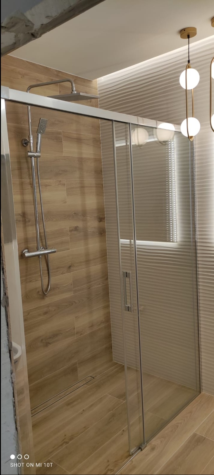Nowoczesna kabina prysznicowa z przesuwnymi drzwiami, wykończona płytkami imitującymi drewno, z odpływem liniowym i designerską lampą wiszącą.