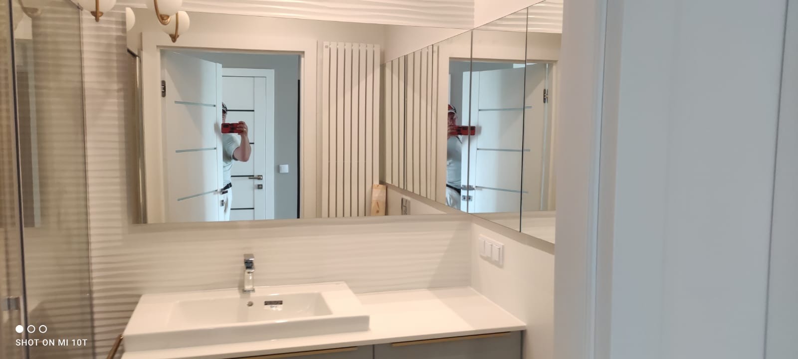 Nowoczesna łazienka z białymi płytkami imitującymi fale, umywalką nablatową i dużym lustrem, w którym odbija się osoba robiąca zdjęcie telefonem.