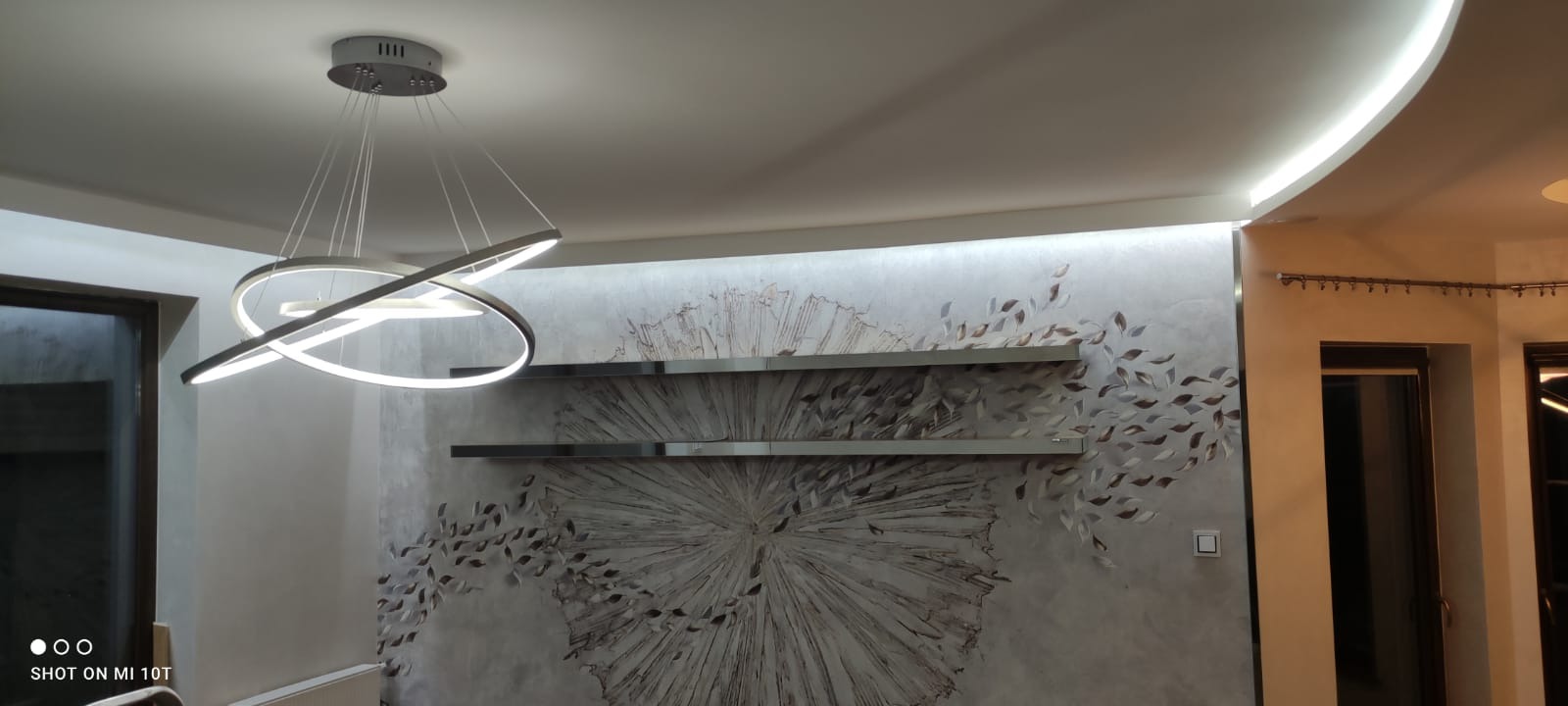 Nowoczesne oświetlenie sufitowe w kształcie pierścieni nad ścianą z dekoracyjną fakturą i metalowymi półkami.