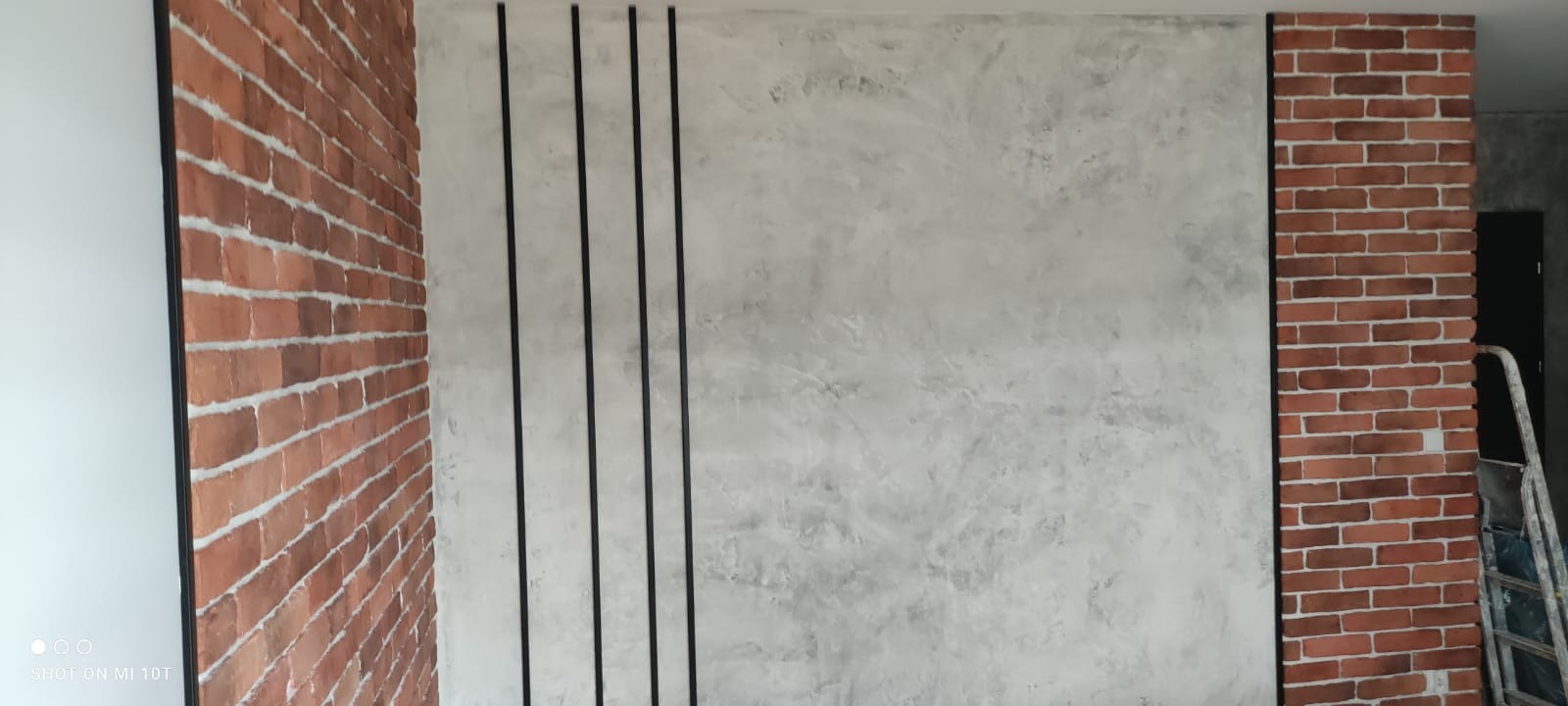 Ściana z efektem betonu architektonicznego, ozdobnymi czarnymi listwami i fragmentami muru z cegły, widoczna drabina malarska w tle.