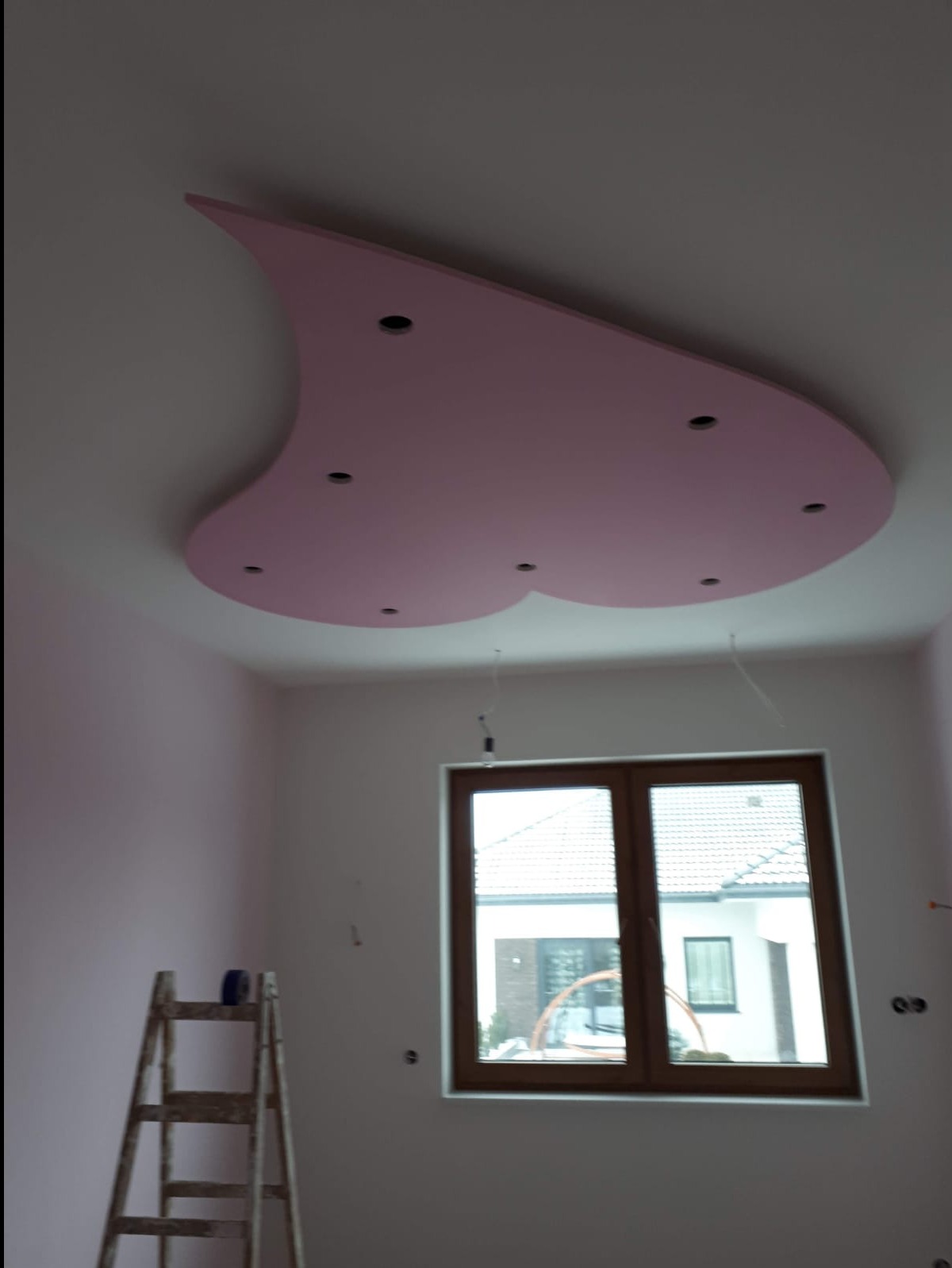 Różowy sufit podwieszany z płyt karton gips o nieregularnym kształcie z otworami na oświetlenie punktowe, widok na okno i drabinę w pokoju podczas remontu.