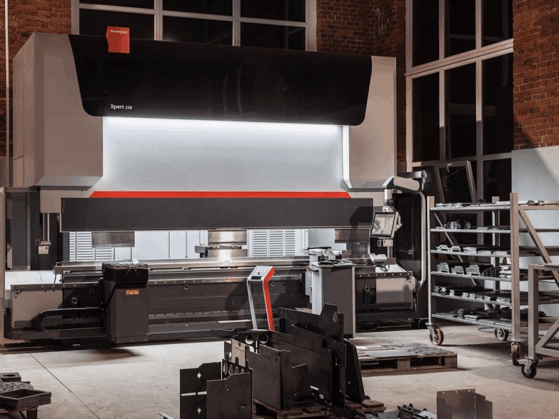 Duża, nowoczesna prasa krawędziowa Xpert 250 w hali produkcyjnej z ceglaną ścianą w tle. Na pierwszym planie widoczne metalowe elementy czekające na obróbkę.