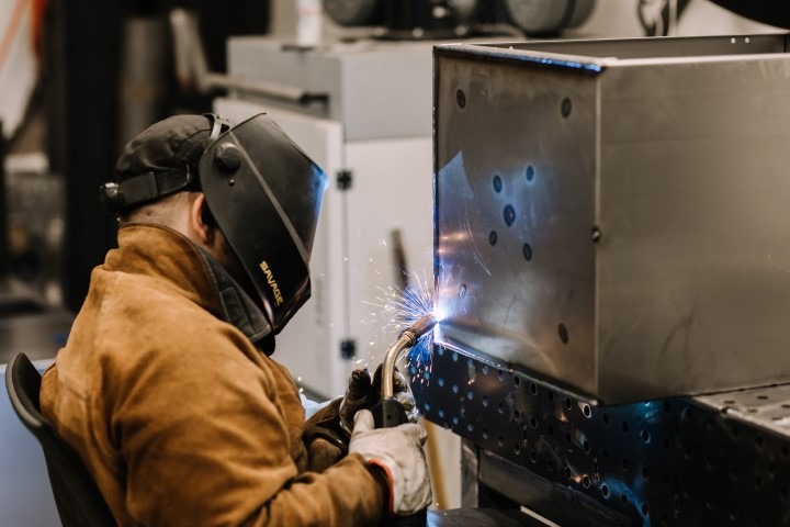 Spawacz w brązowej kurtce i masce ochronnej marki Savage, spawający metalową skrzynkę z widocznymi iskrami i otworami na śruby.