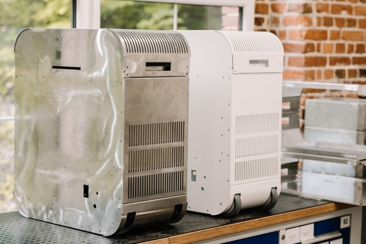 Dwa metalowe urządzenia na kółkach, jeden surowy srebrny, drugi pomalowany na biało, stoją obok siebie na stole warsztatowym w pomieszczeniu z ceglaną ścianą w tle.