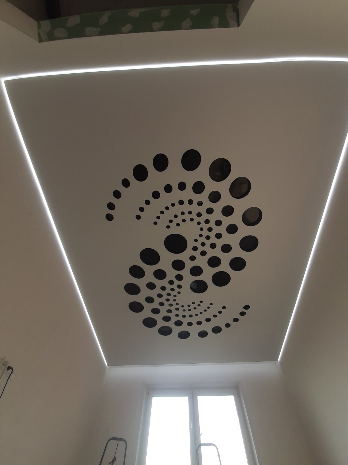 Biały sufit napinany z czarnym, spiralnym wzorem z kropek i oświetleniem LED po obwodzie. Widok z dołu na okno i fragment drabiny.
