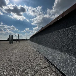 Nowa papa na płaskim dachu pokryta posypką mineralną, widoczne kominy wentylacyjne, błękitne niebo z chmurami i fragment dachu sąsiedniego budynku.