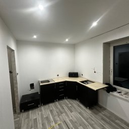 Maksymilian Dulewski - Wnętrze w trakcie prac wykończeniowych: czarne szafki kuchenne z beżowymi blatami zamontowane w pomieszczeniu z szarą podłogą imitującą drewno, widoczne niedokończone instalacje elektryczne...