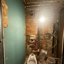 Maksymilian Dulewski - Łazienka w trakcie prac: widoczna ściana z płyt kartonowo-gipsowych, odsłonięta cegła, rury, kable, surowy strop, toaleta i pojemniki z zaprawą.