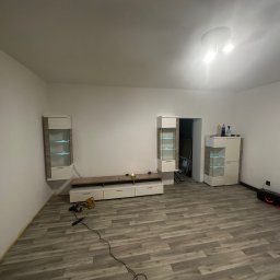 Maksymilian Dulewski - Pokój z nowymi panelami podłogowymi w odcieniach szarości, meblami w kolorze białym i beżowym oraz widocznymi narzędziami do montażu podłogi.