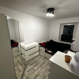 Maksymilian Dulewski - Widok na umeblowane wnętrze pokoju z łóżkiem, komodami i szafą z lustrzanymi drzwiami, oświetlone trzypunktową lampą sufitową.