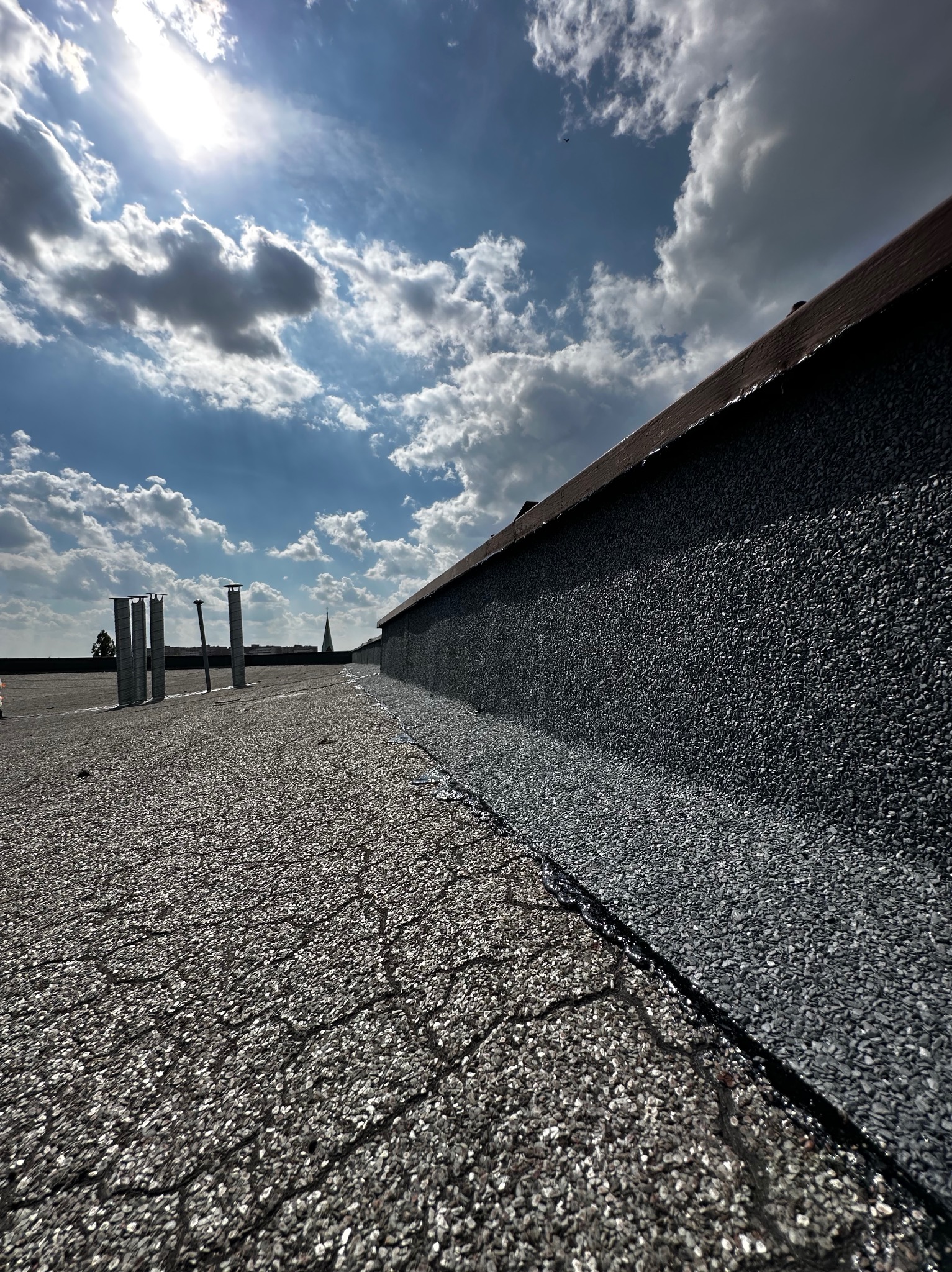 Nowa papa na płaskim dachu pokryta posypką mineralną, widoczne kominy wentylacyjne, błękitne niebo z chmurami i fragment dachu sąsiedniego budynku.