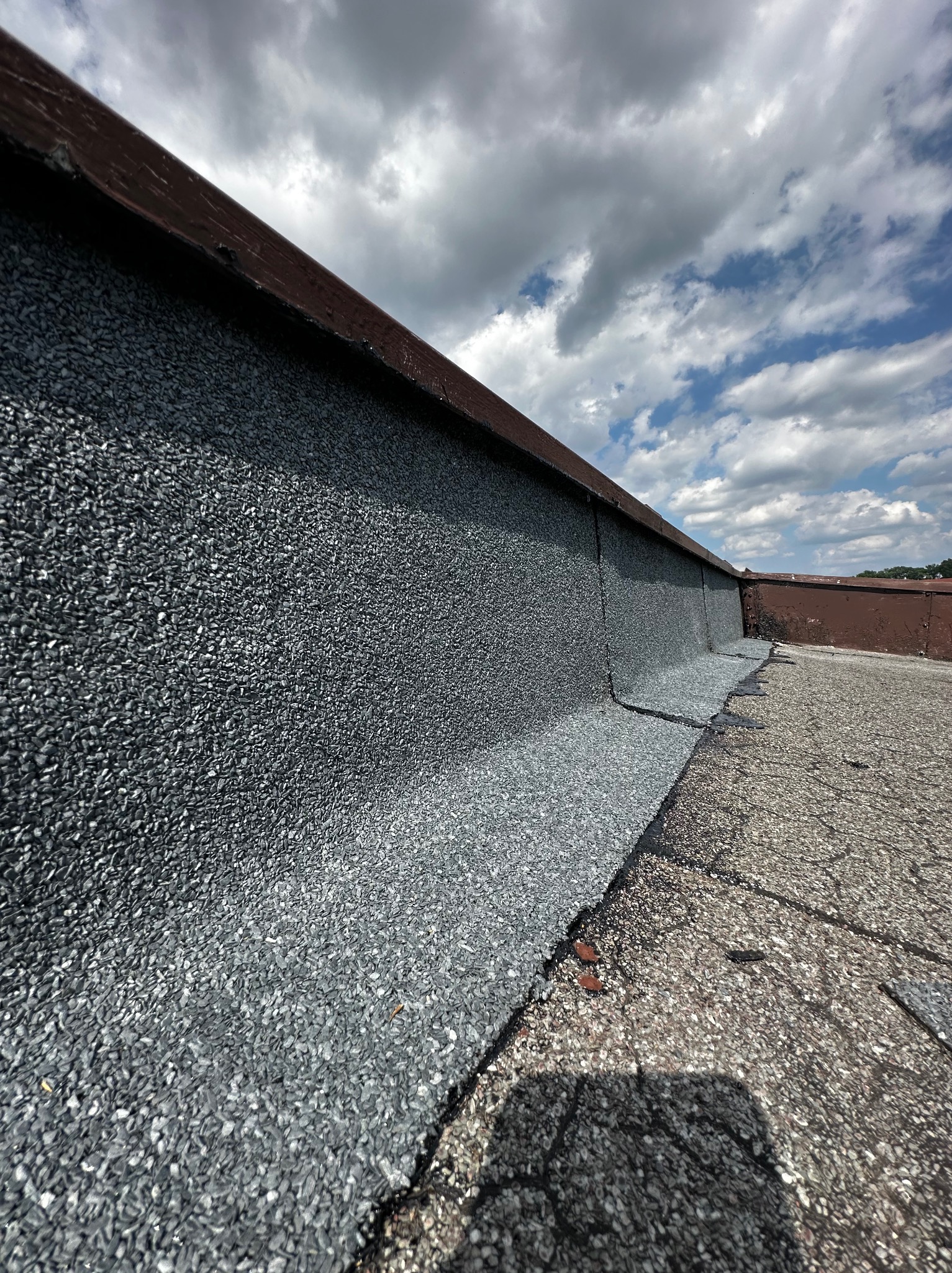 Szczegółowe ujęcie krawędzi dachu pokrytej szarą papą z posypką mineralną, z widoczną metalową obróbką blacharską i fragmentem starego pokrycia, na tle pochmurnego nieba.