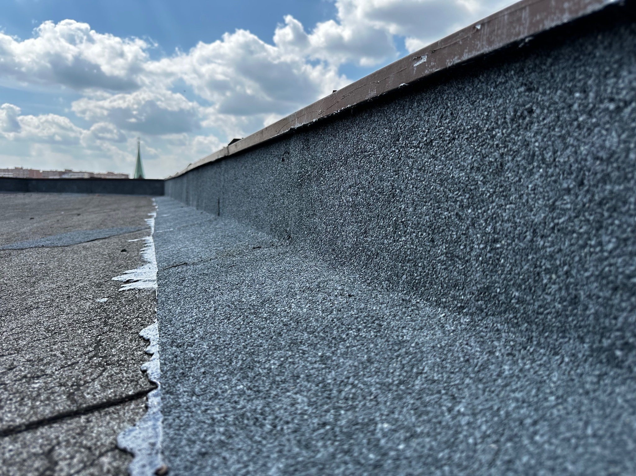 Szczegółowe ujęcie powierzchni dachu pokrytej papą z drobnym granulatem, widoczne połączenie z obróbką blacharską i fragment panoramy miasta w tle pod błękitnym niebem.