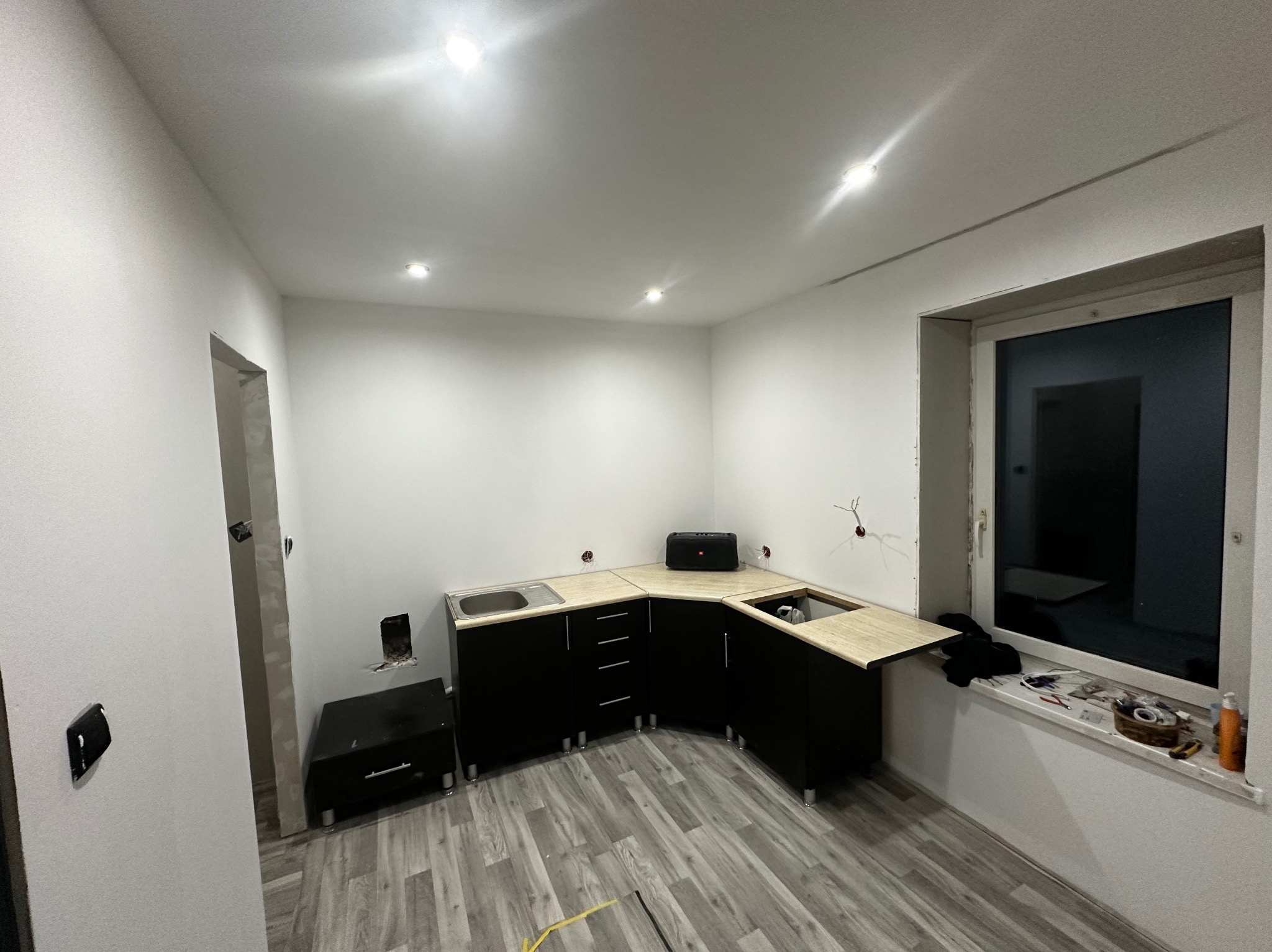 Wnętrze w trakcie prac wykończeniowych: czarne szafki kuchenne z beżowymi blatami zamontowane w pomieszczeniu z szarą podłogą imitującą drewno, widoczne niedokończone instalacje elektryczne...