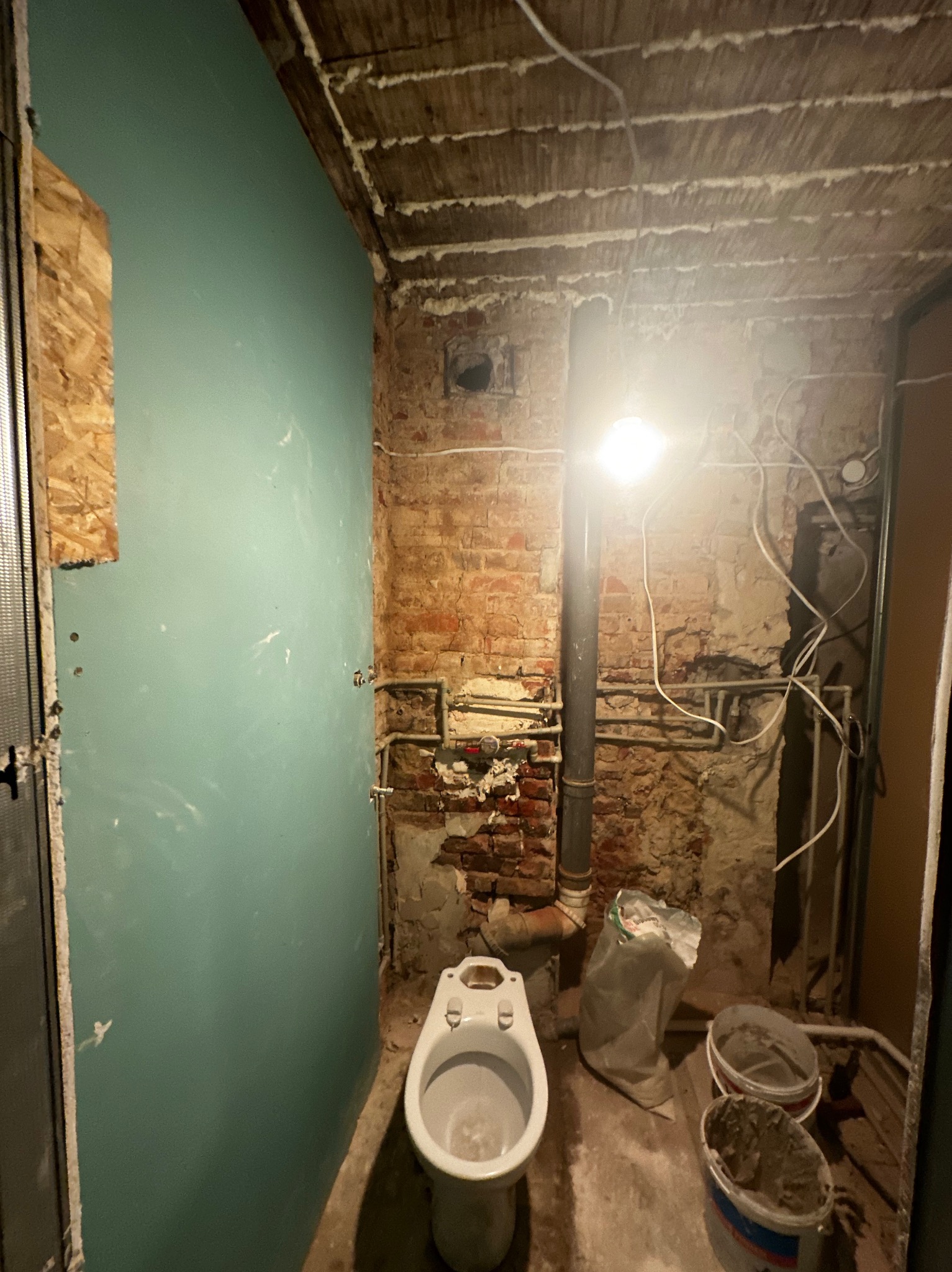 Łazienka w trakcie prac: widoczna ściana z płyt kartonowo-gipsowych, odsłonięta cegła, rury, kable, surowy strop, toaleta i pojemniki z zaprawą.