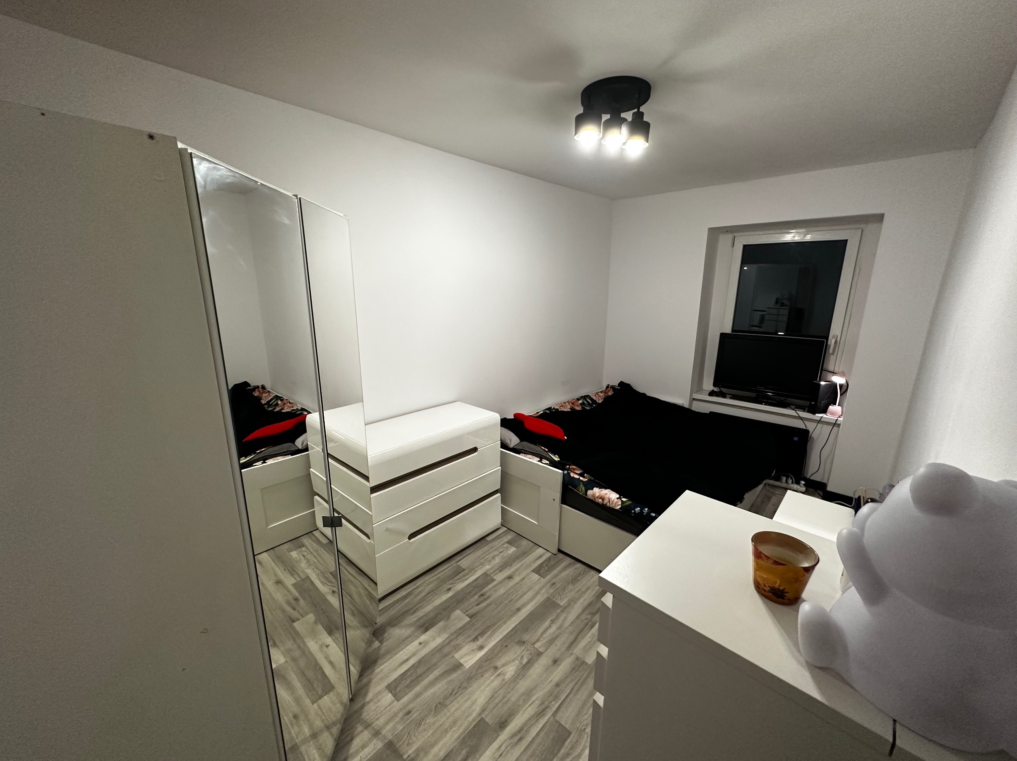 Widok na umeblowane wnętrze pokoju z łóżkiem, komodami i szafą z lustrzanymi drzwiami, oświetlone trzypunktową lampą sufitową.