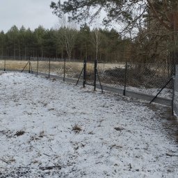 Maciej Budpol - Długa siatka ogrodzeniowa z betonową podmurówką, widok na zaśnieżone pole i las w tle, pochmurny dzień.