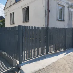 Szare, metalowe ogrodzenie palisadowe z bramą wjazdową na posesję z budynkiem w tle, widoczne ślady świeżego betonu u podstawy.
