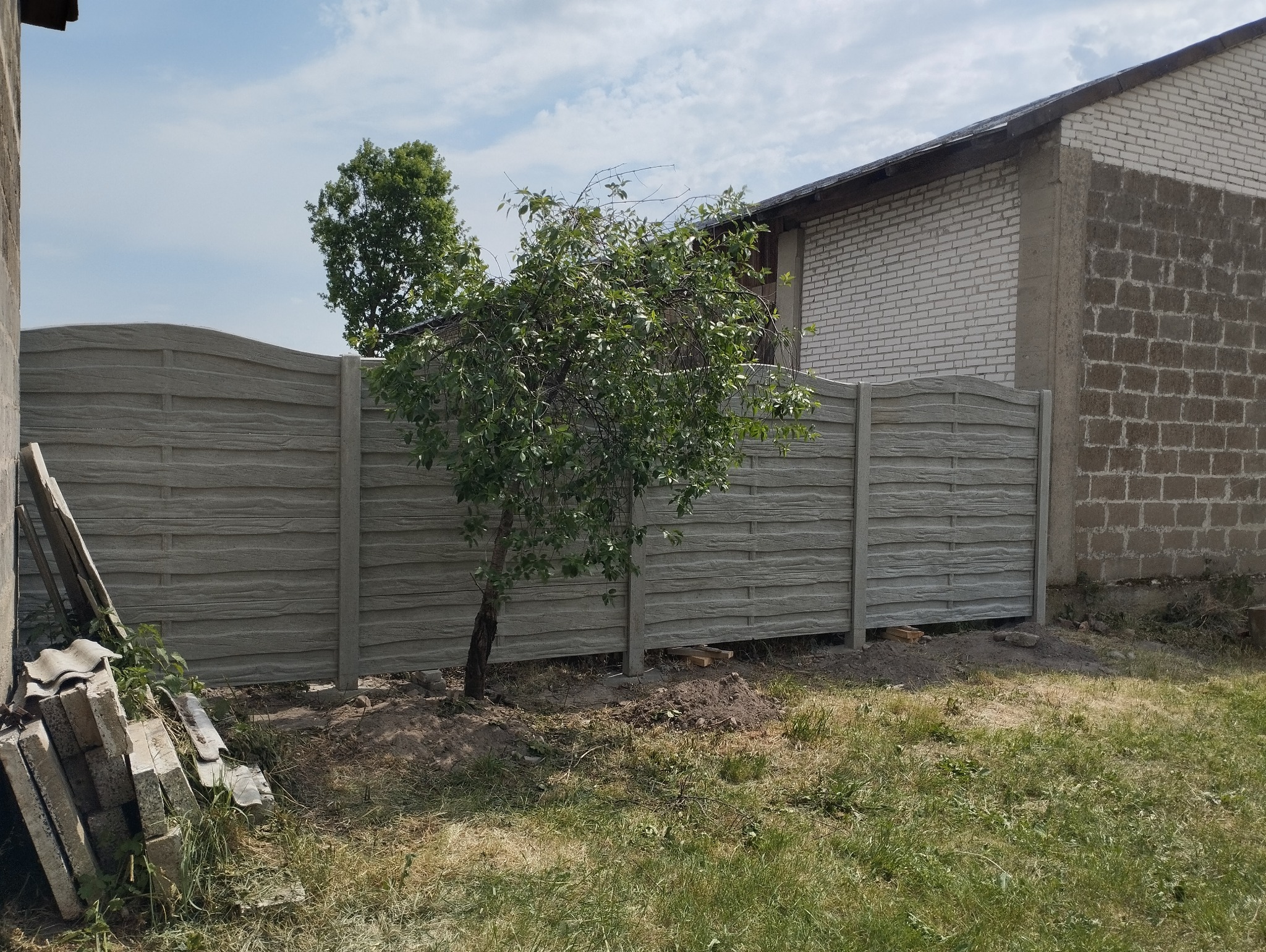 Szary, betonowy płot z poziomymi panelami imitującymi drewno, na tle trawnika i budynku z cegły, z małym drzewkiem rosnącym przed płotem. Na pierwszym planie stos starych dachówek.