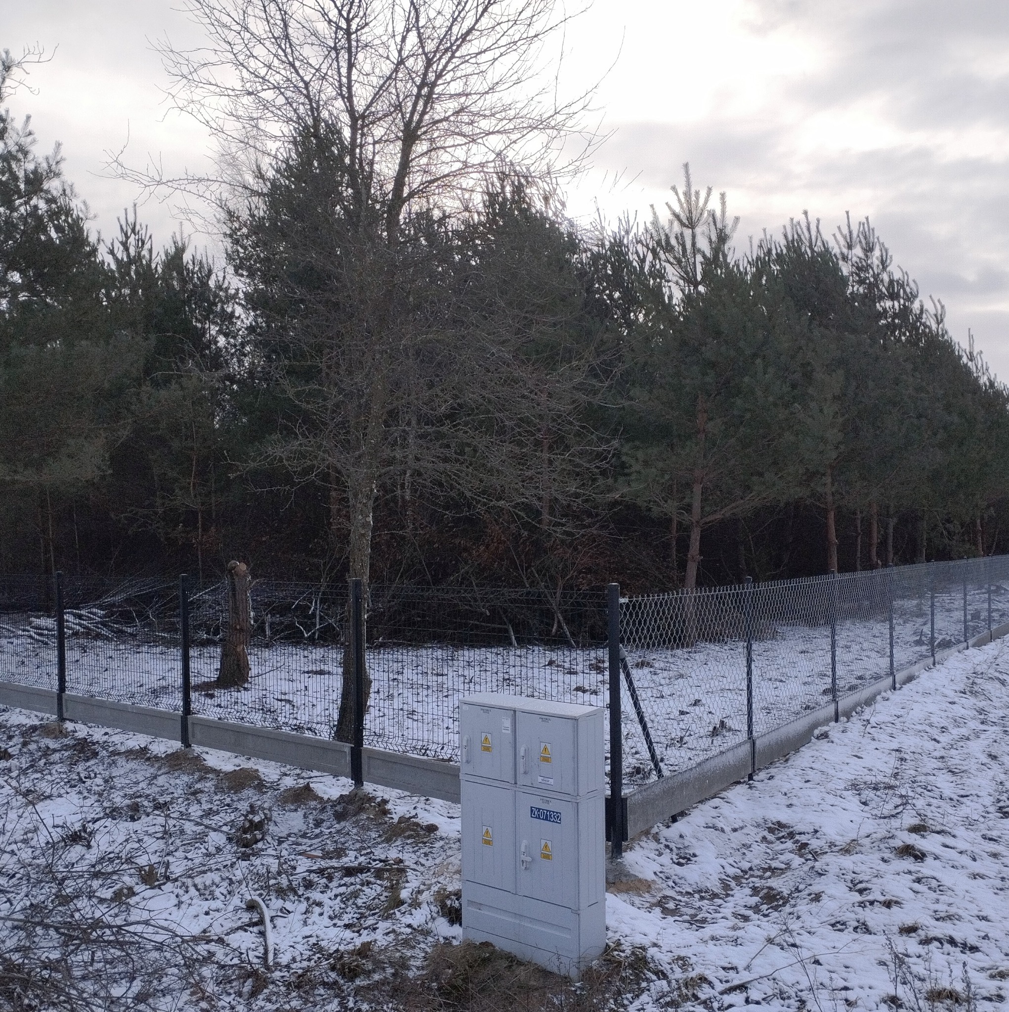 Fragment posesji z siatką ogrodzeniową na betonowej podmurówce, widoczna skrzynka elektryczna i zasypany śniegiem teren z drzewami w tle.