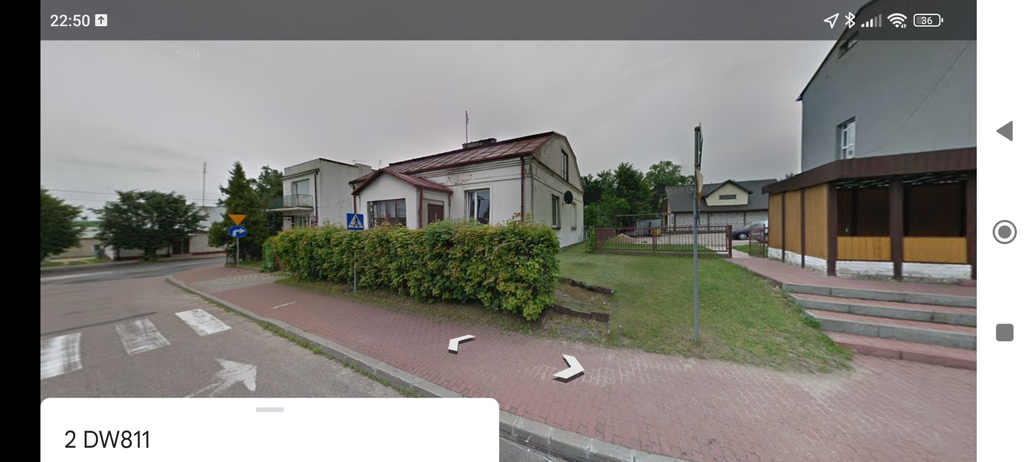 Widok z ulicy na dom z ogrodzeniem z żywopłotu i znakami drogowymi, z widocznym fragmentem chodnika i jezdni.