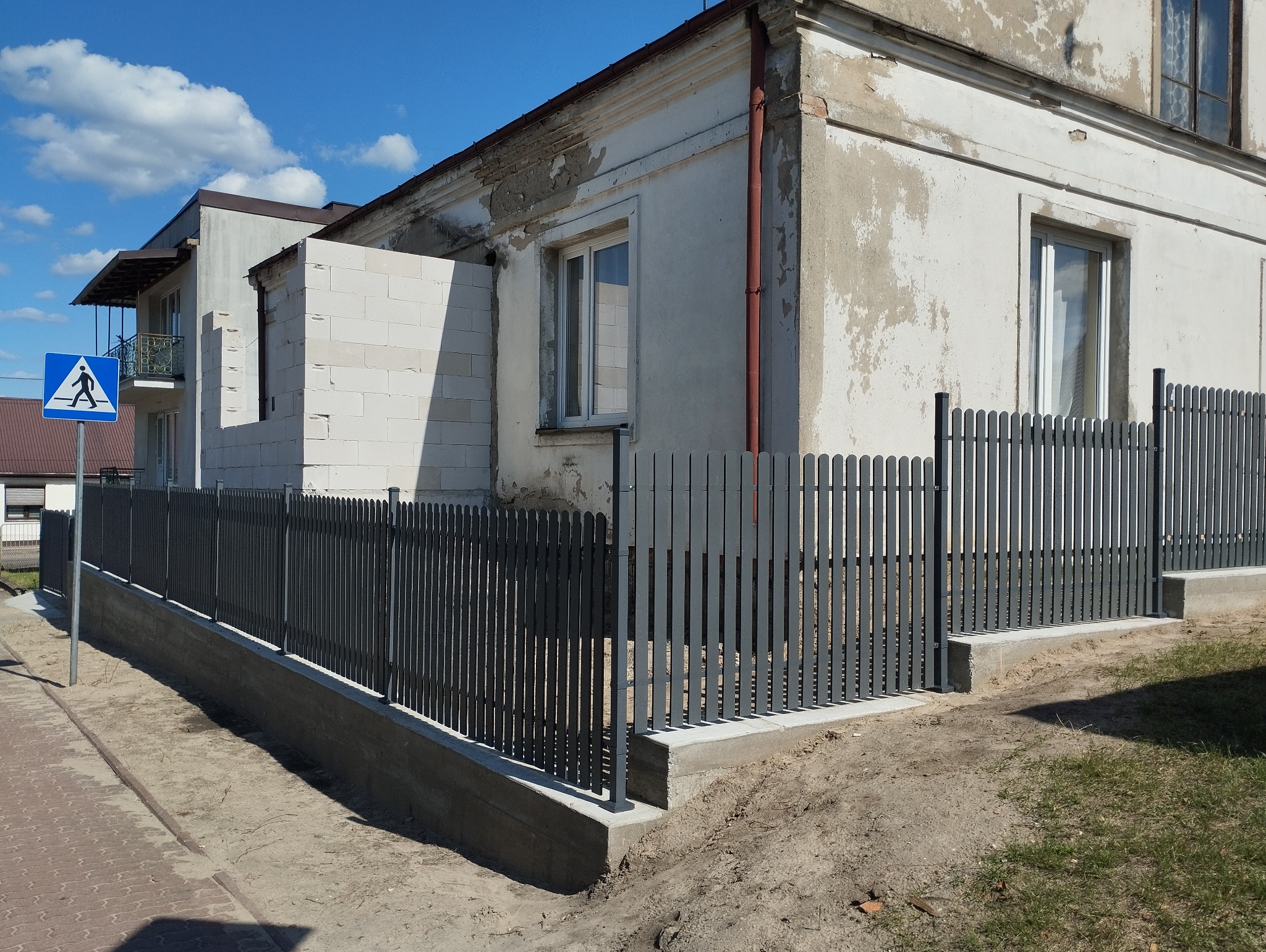 Nowoczesne, szare ogrodzenie metalowe typu sztachetowego na betonowej podmurówce, widoczne na tle budynku w stanie surowym i znaku drogowego.