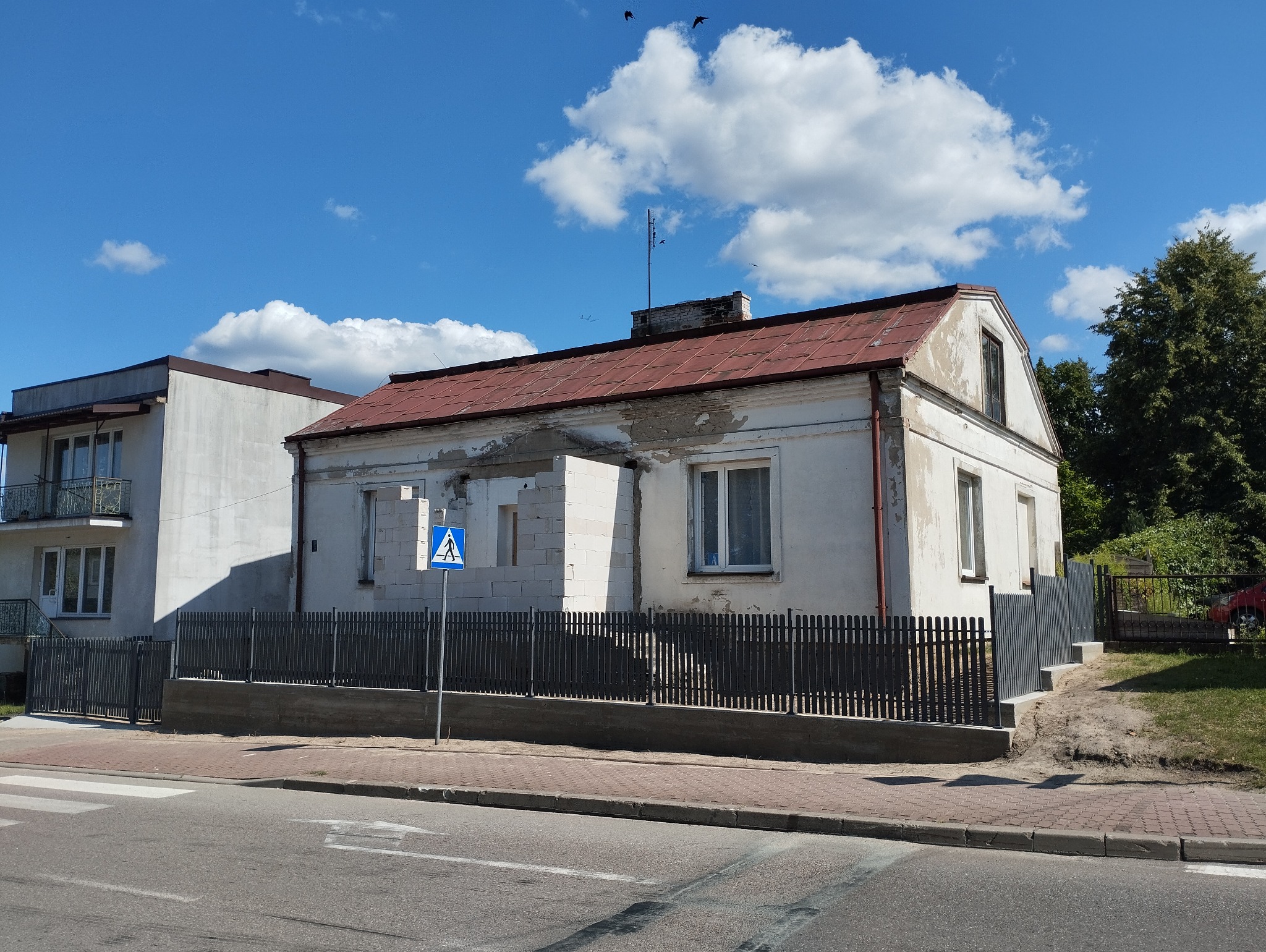 Szary płot sztachetowy przed domem w trakcie przebudowy, widoczny fragment dobudówki z białego bloczka, znak przejścia dla pieszych, błękitne niebo z chmurami.