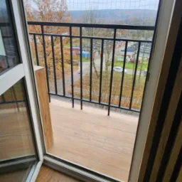 Balkon z czarną, metalową balustradą, zabezpieczony siatką, z widokiem na jesienny krajobraz miejski.