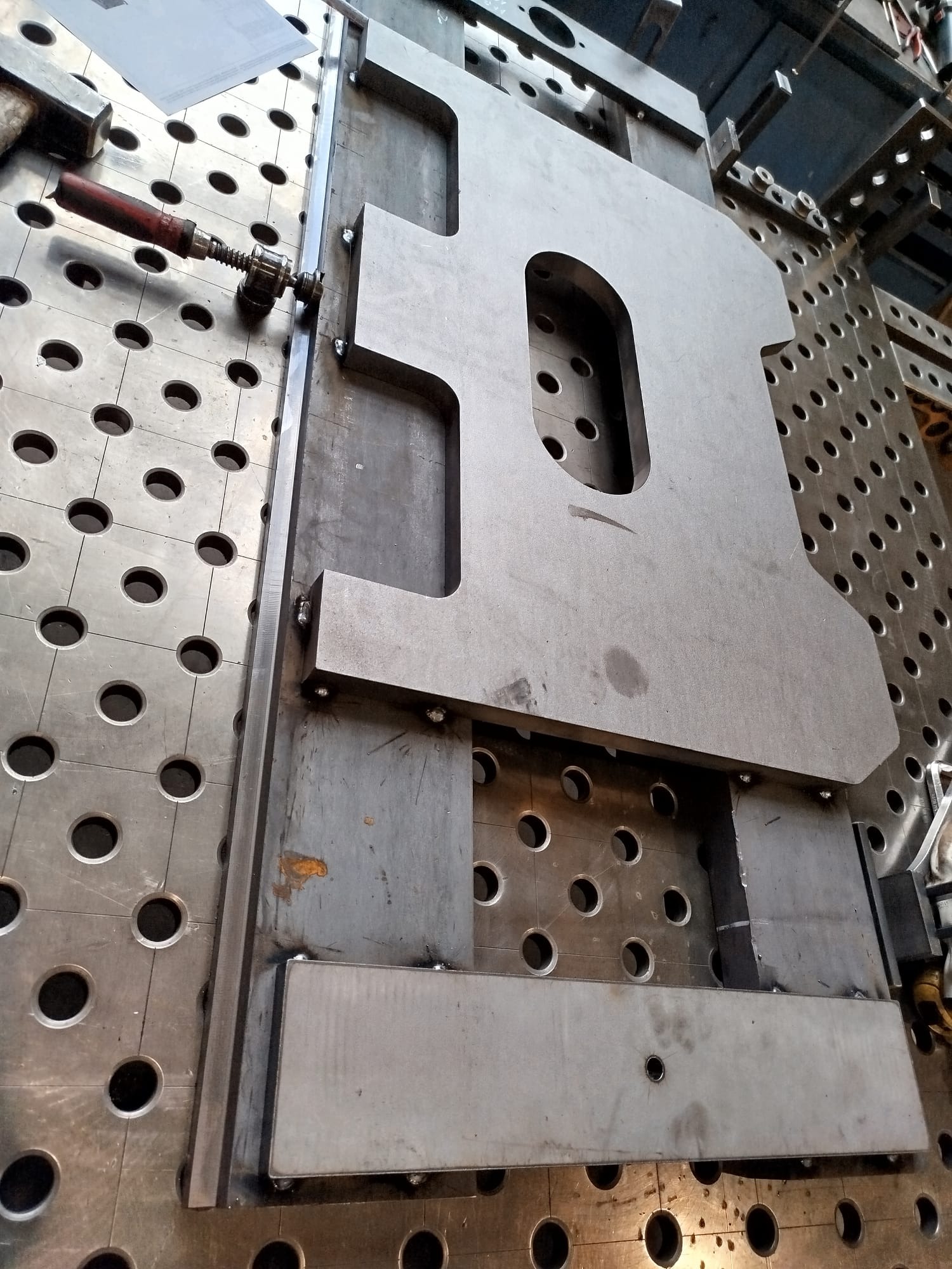 Metalowa płyta z wyciętym otworem, mocowana do perforowanego stołu spawalniczego za pomocą ścisku stolarskiego, widoczne ślady spawania.