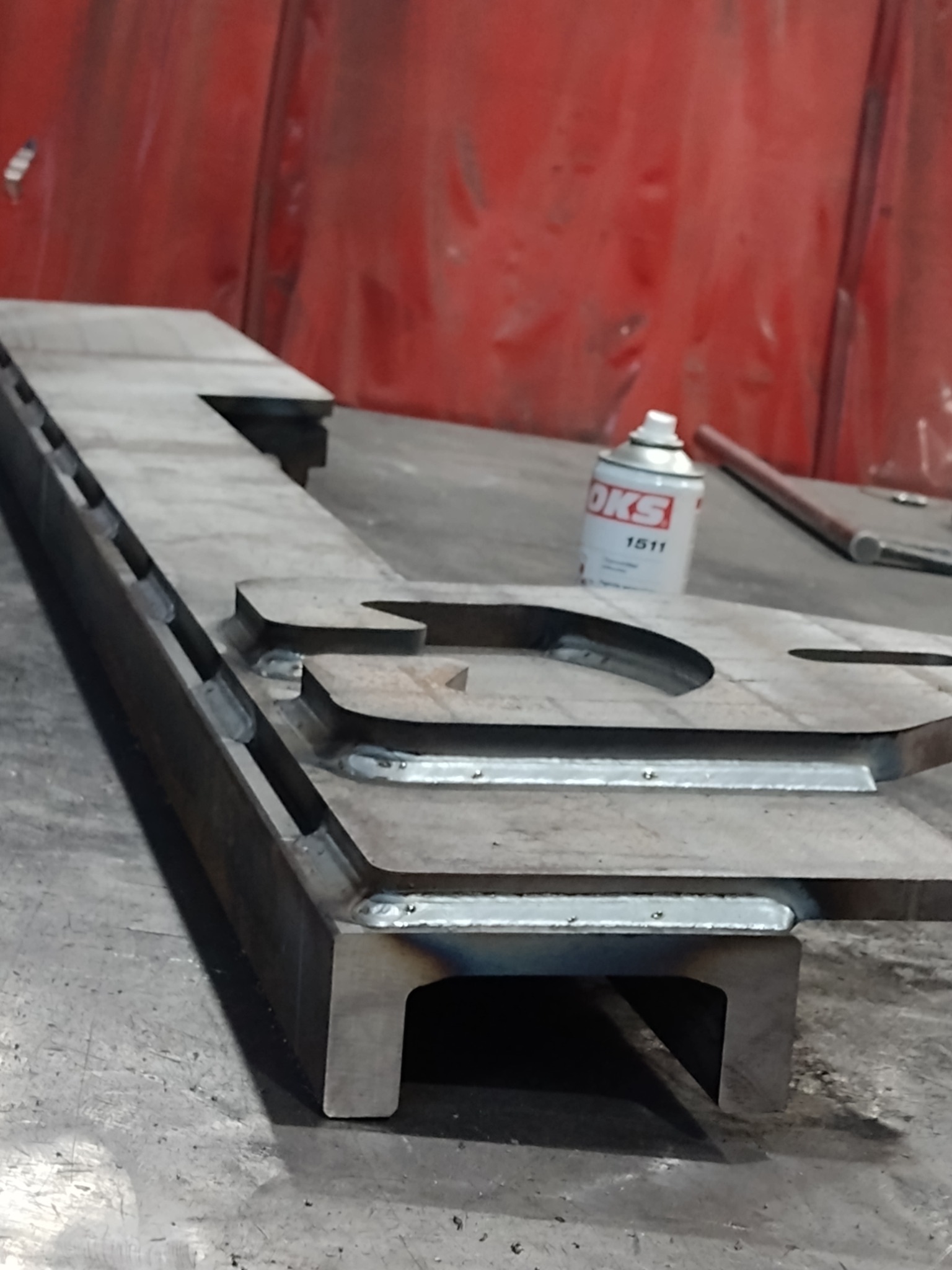 Metalowy element konstrukcyjny z widocznymi spawami, leżący na stole warsztatowym, obok puszka ze smarem w tle czerwona ściana warsztatu.