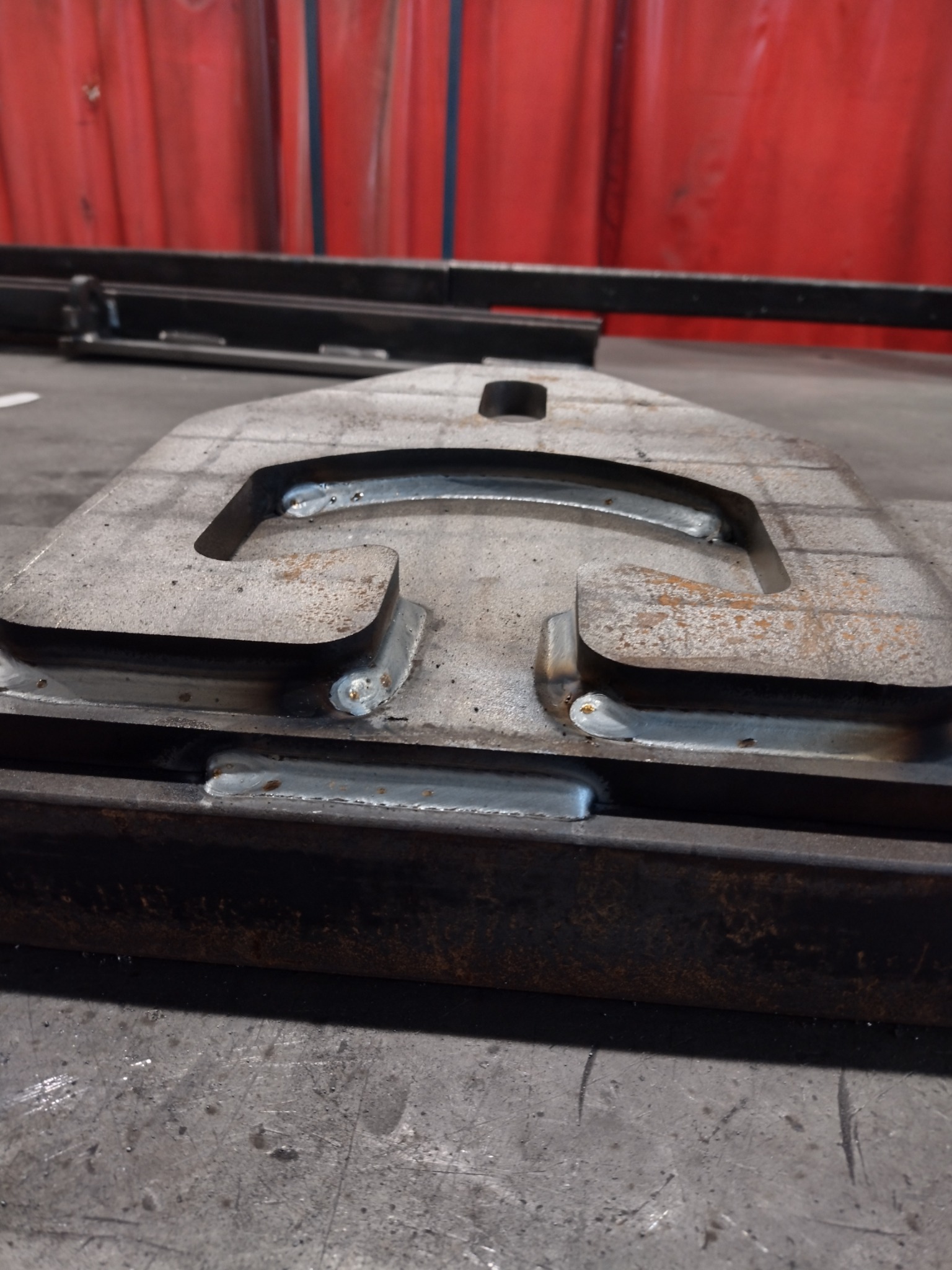 Metalowy element z widocznymi spawami, leżący na metalowej konstrukcji, z czerwonym materiałem w tle.