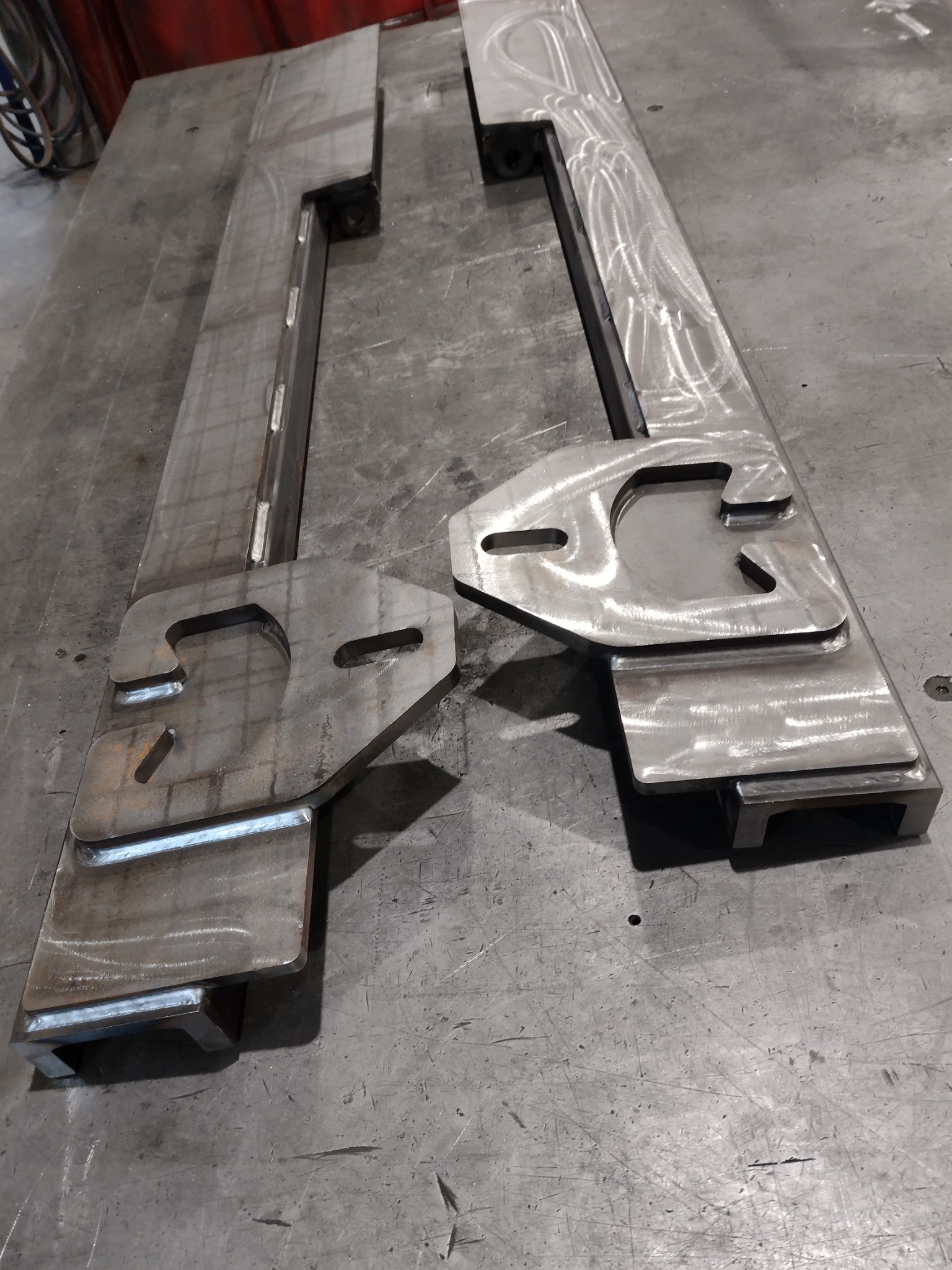Dwie metalowe konstrukcje z przyspawanymi elementami, leżące na metalowym stole, widoczne ślady obróbki i spoiny