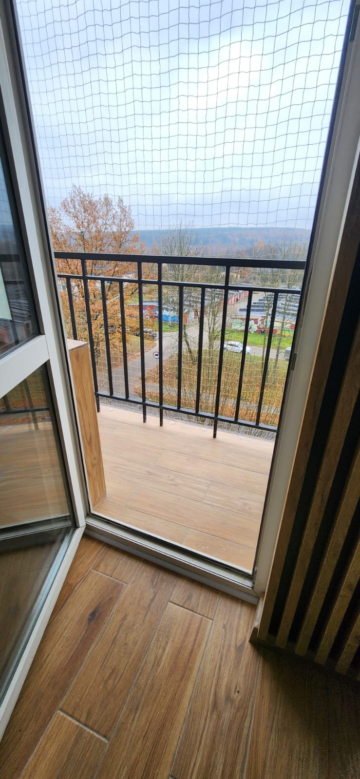 Balkon z czarną, metalową balustradą, zabezpieczony siatką, z widokiem na jesienny krajobraz miejski.