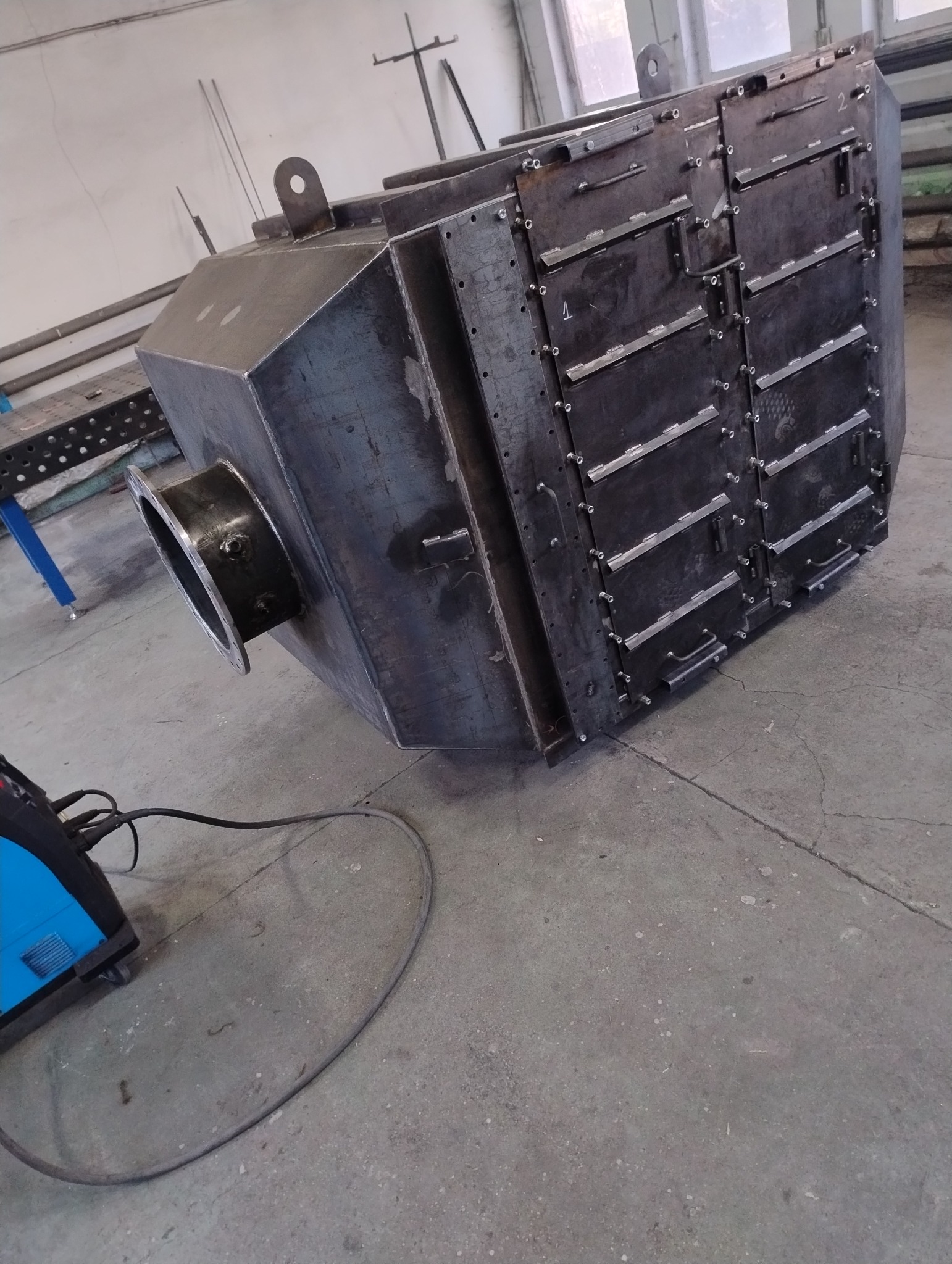 Spawana, surowa konstrukcja metalowa z okrągłym otworem i uchwytem, widoczna spawarka obok, w warsztacie.