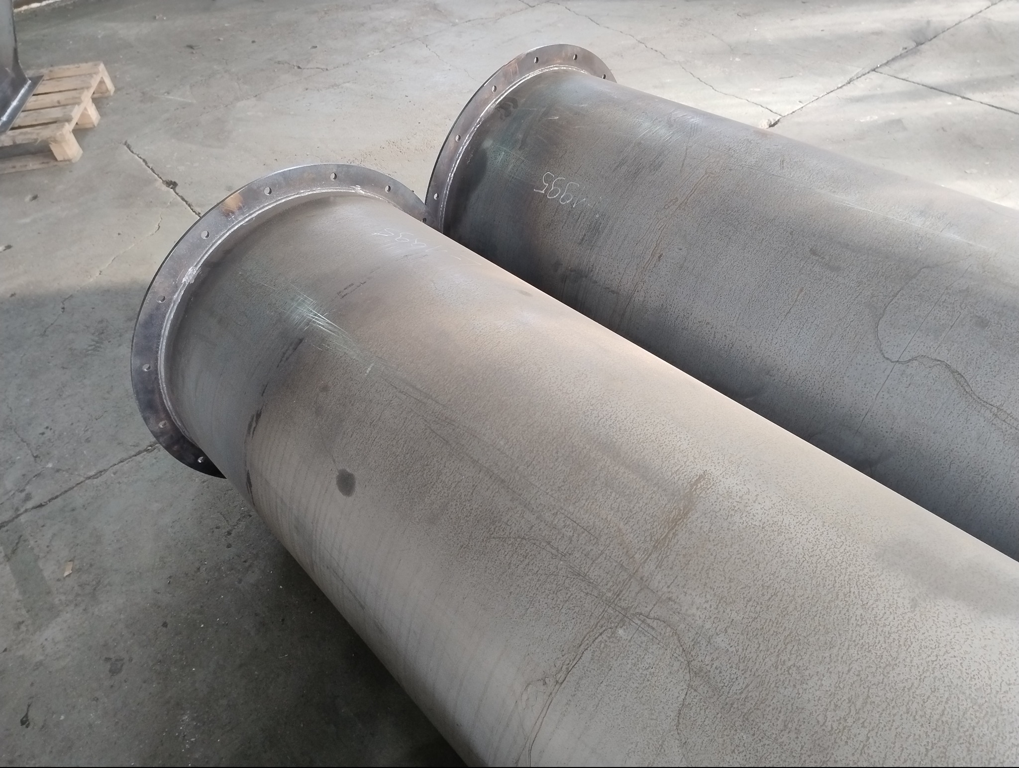 Dwa duże, stalowe cylindryczne elementy z przyspawanymi kołnierzami, leżące na betonowej podłodze. Na jednym z cylindrów widoczne białe napisy.