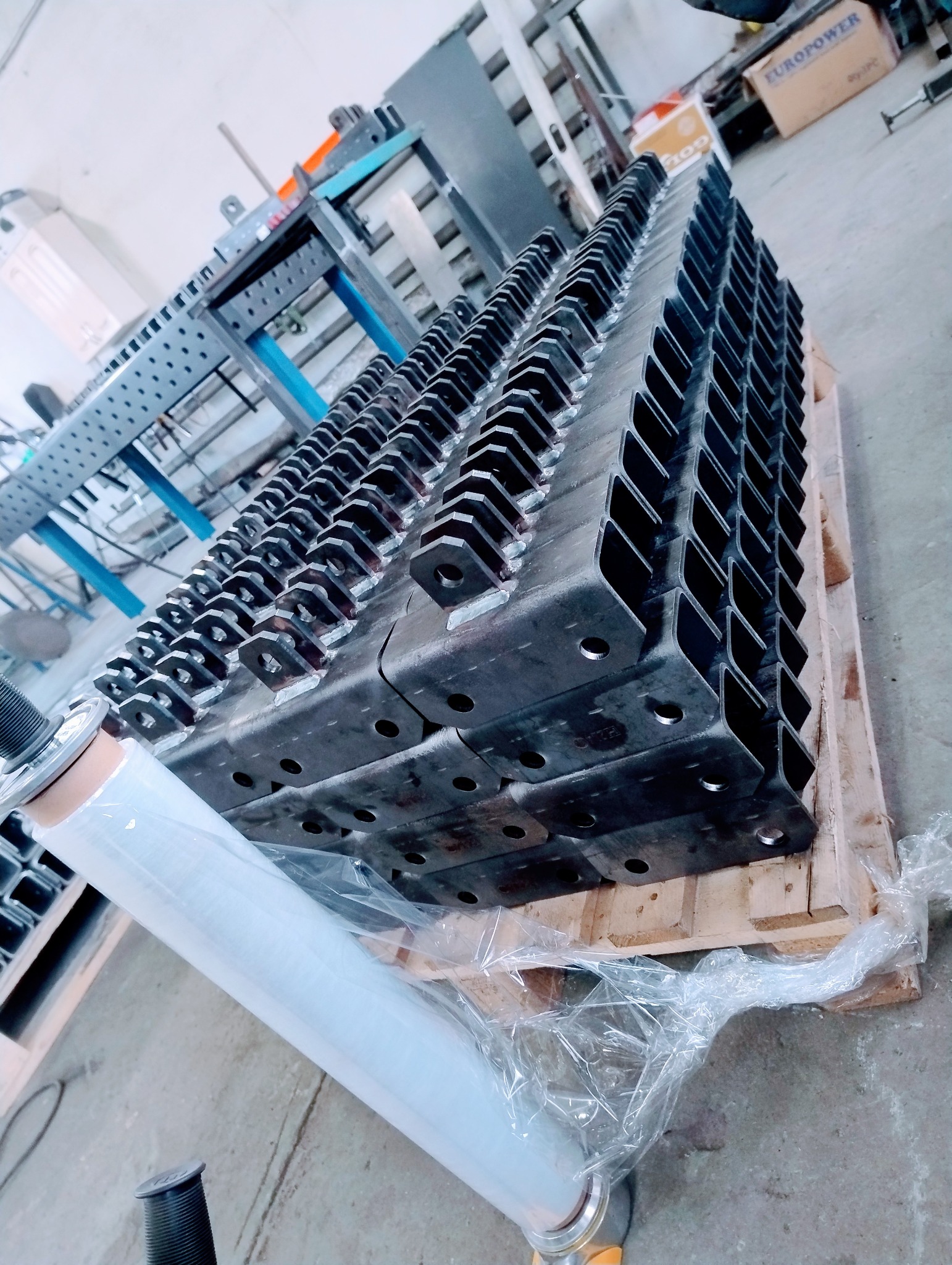 Stos metalowych wsporników z otworami, ułożonych na palecie i owiniętych folią stretch, widoczny fragment stołu spawalniczego w tle.