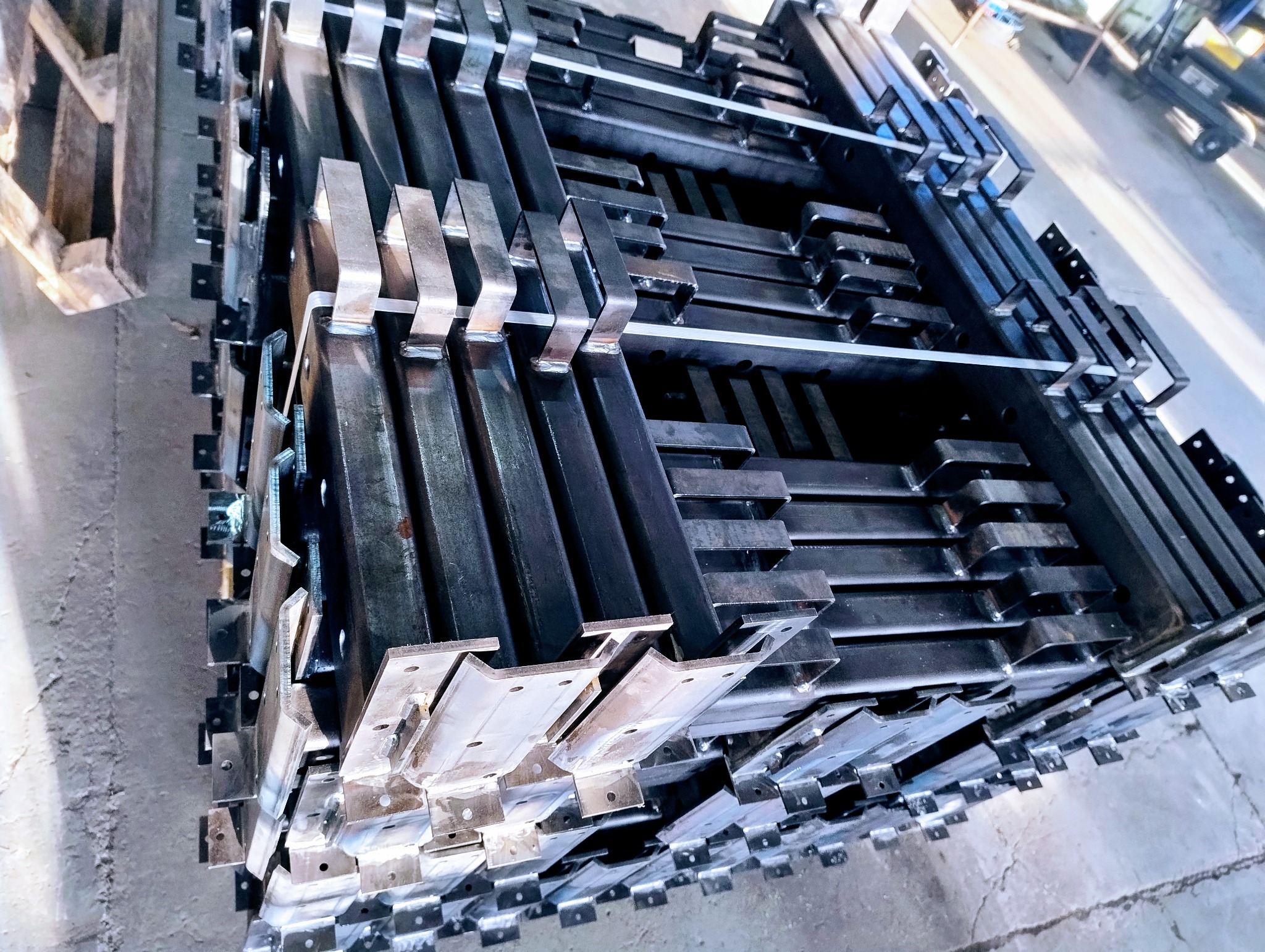 Stos metalowych elementów konstrukcyjnych z przyspawanymi uchwytami, zabezpieczonych białymi taśmami, na szarym betonowym podłożu.