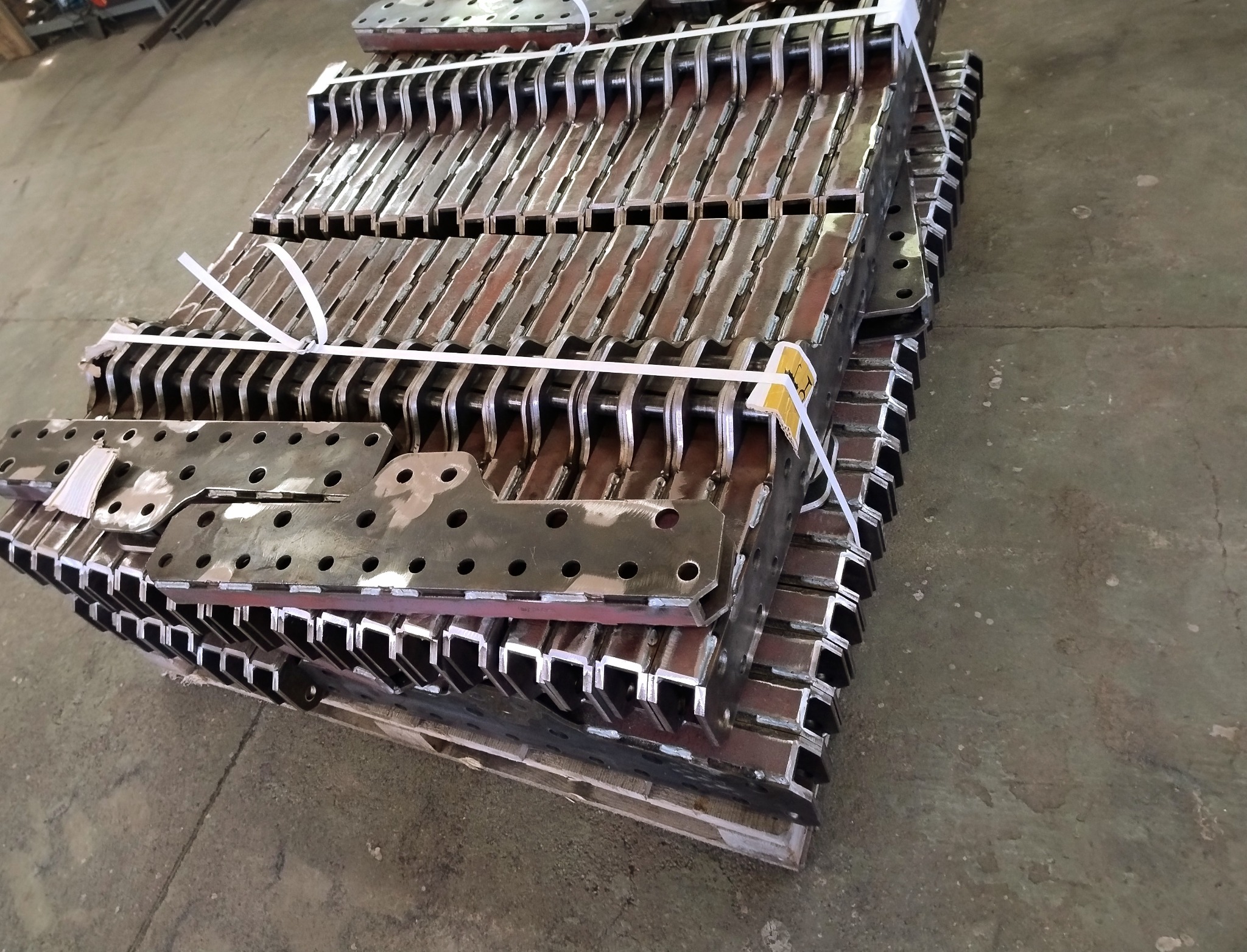 Stos metalowych elementów z otworami, połączonych białym sznurkiem na palecie. Widoczne spawy i surowa powierzchnia stali, sugerujące etap produkcji.