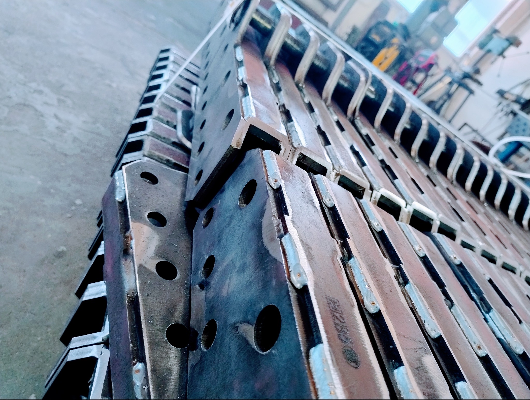 Stos metalowych elementów z otworami, połączonych spawami. Elementy ustawione pionowo, widoczne ślady obróbki. W tle warsztat.