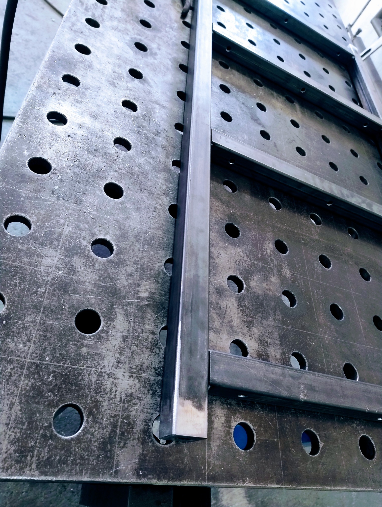 Metalowa konstrukcja spawana na perforowanym stole spawalniczym. Widoczny fragment ramy z profili zamkniętych, surowe wykończenie, detale spawów.