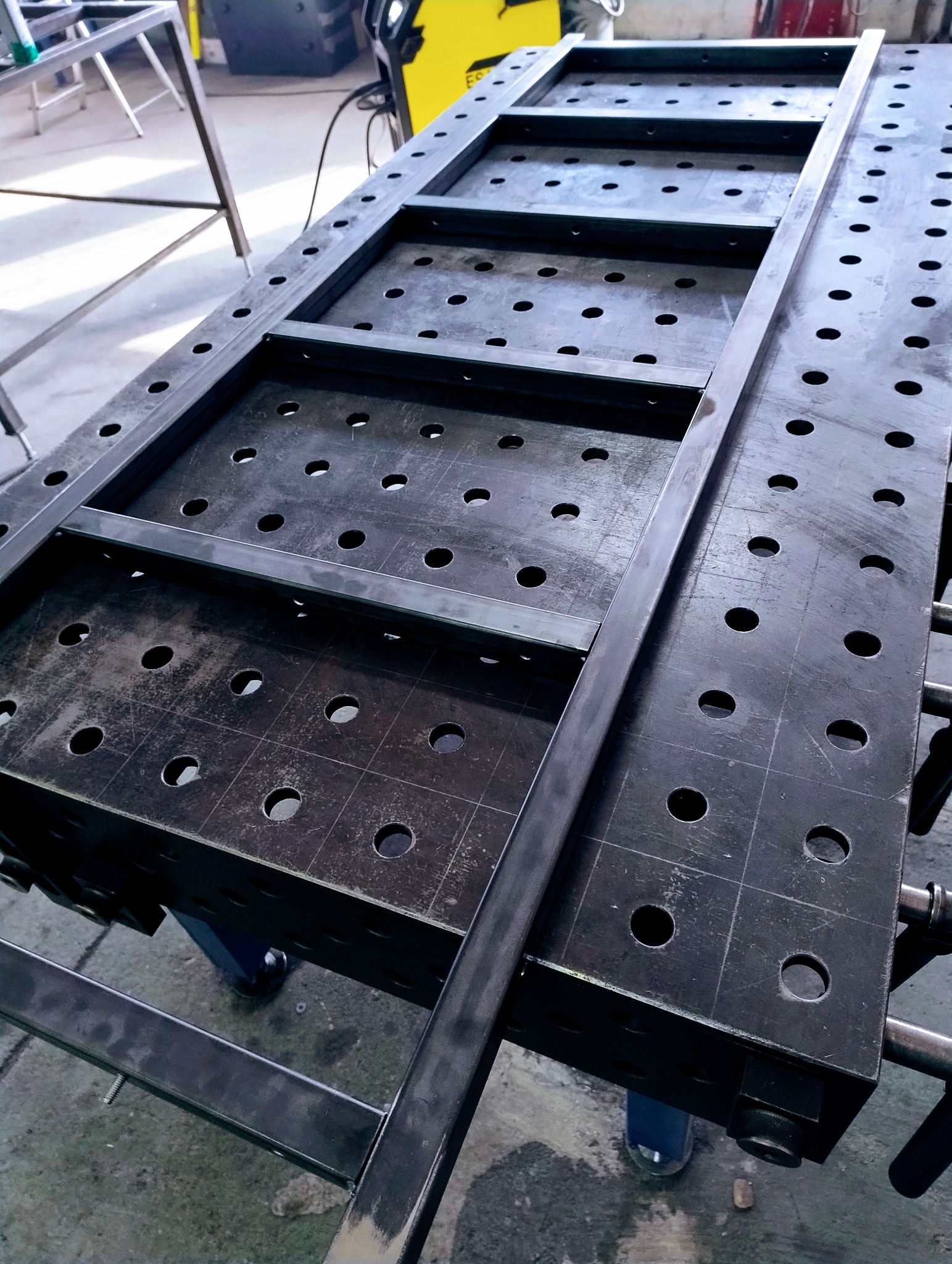 Konstrukcja metalowa z profili na perforowanym stole spawalniczym, przygotowana do spawania. Widoczny spawacz w tle.