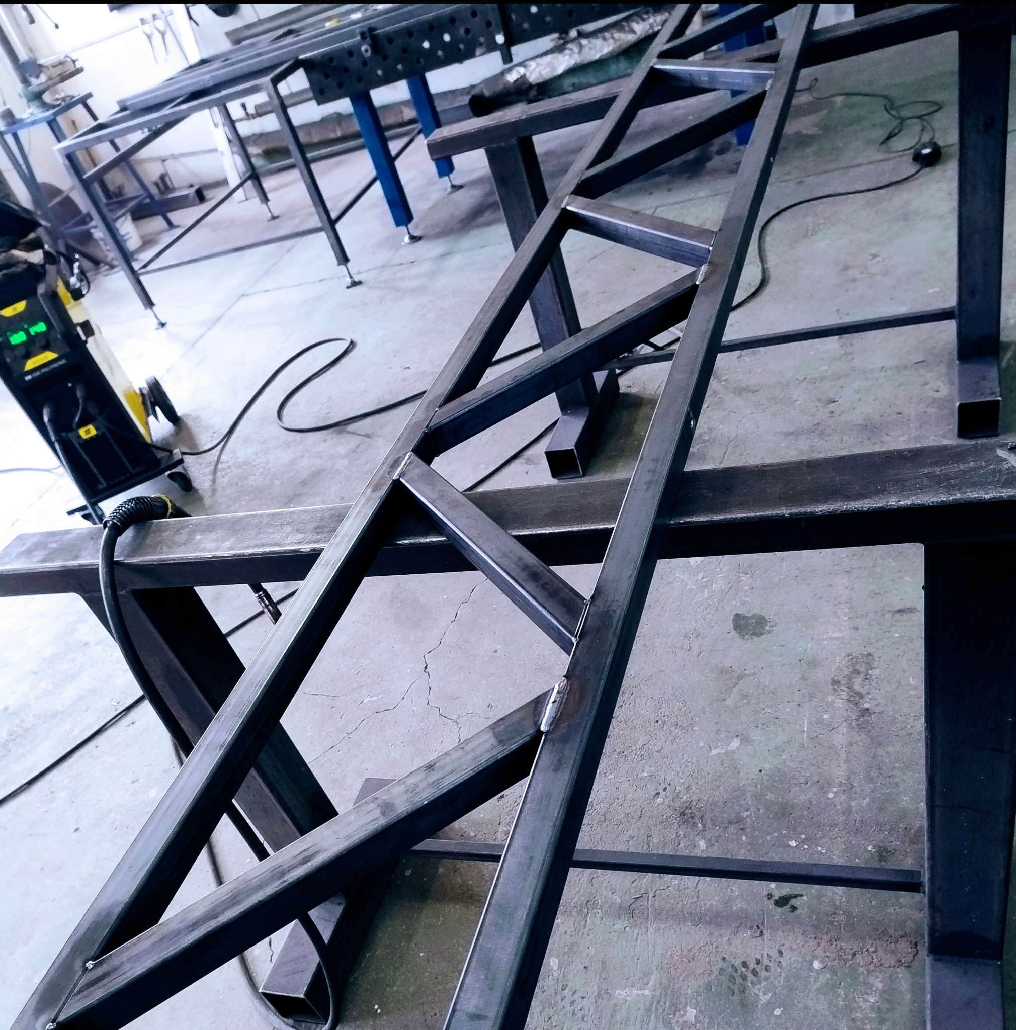 Metalowa konstrukcja spawana, z widocznymi spawami, na metalowym stole warsztatowym. W tle spawarka i inne stoły warsztatowe.