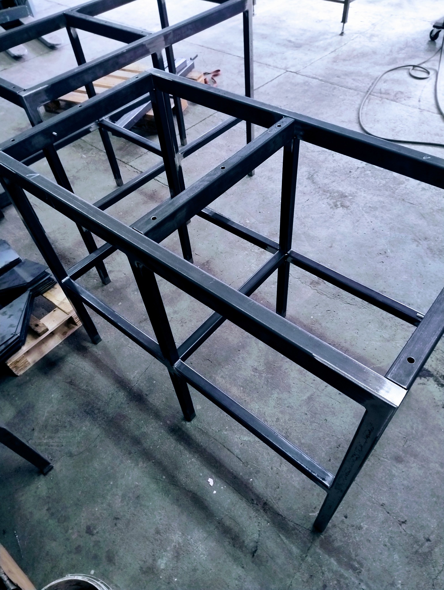 Surowa, spawana konstrukcja metalowa z profili, tworząca szkielet mebla, widoczna w warsztacie na tle betonowej posadzki. Paleta z elementami obok.