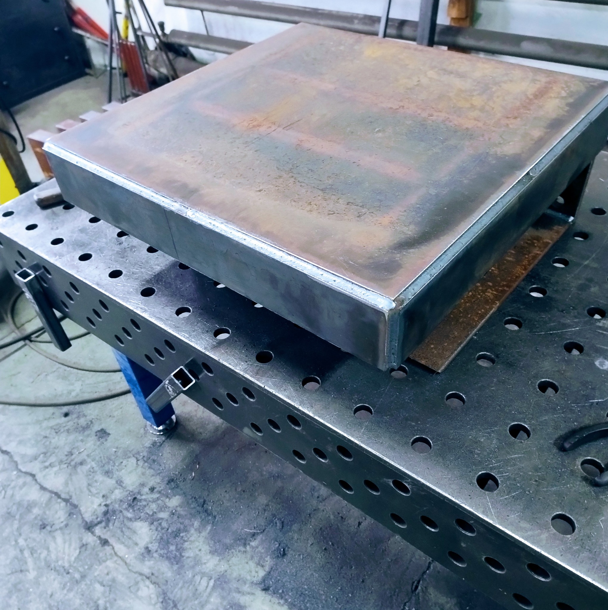 Metalowa konstrukcja spawana, leżąca na perforowanym stole spawalniczym. Widoczne spawy i ślady obróbki metalu. Surowy, industrialny wygląd.