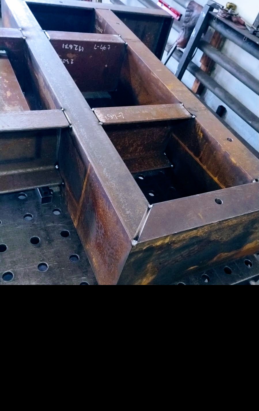 Metalowa konstrukcja spawana na stole spawalniczym z widocznymi spawami i oznaczeniami na elementach. Detal z bliska, ukazujący precyzję wykonania.